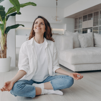 Corperformance - Tile Image - Serene lady sitting crossed legged with hands in meditation pose on the floor of a modern cream living room