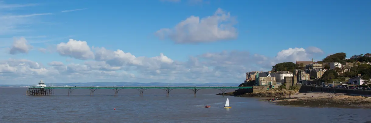 A photo of Clevedon overlooking the pier with bright blue sky, some fluffy clouds and a little sailing boat in the water